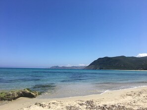 A beach with clear blue water, sandy shore, and a mountain in the background.