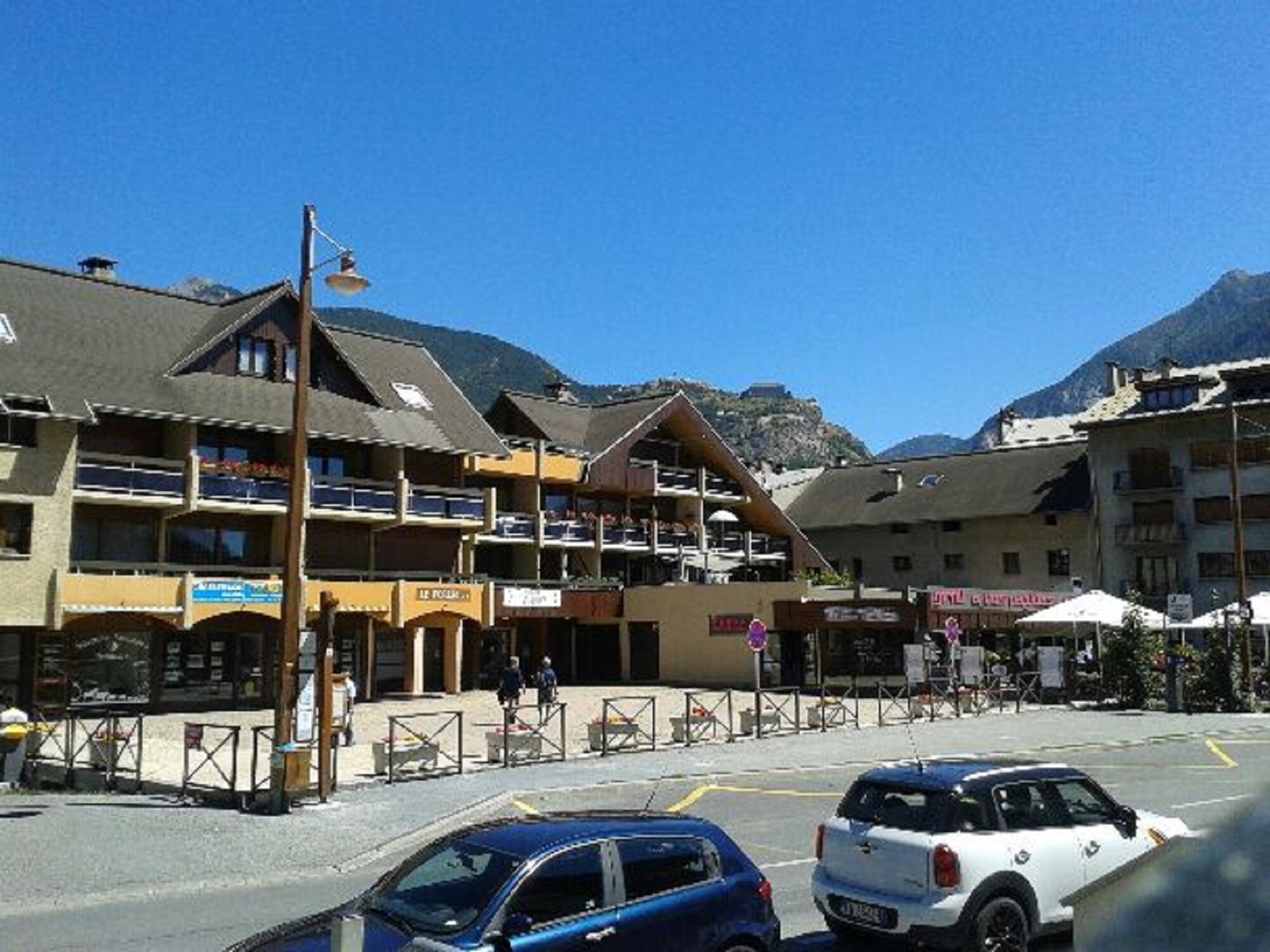 A multi-story building with balconies, a parking area, and mountainous background.
