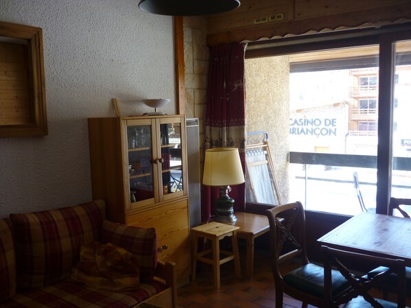 A cozy room with a wooden cabinet, a plaid sofa, a small table with a lamp, and a view of a building with a sign reading "CASINO DE BRIANCON".