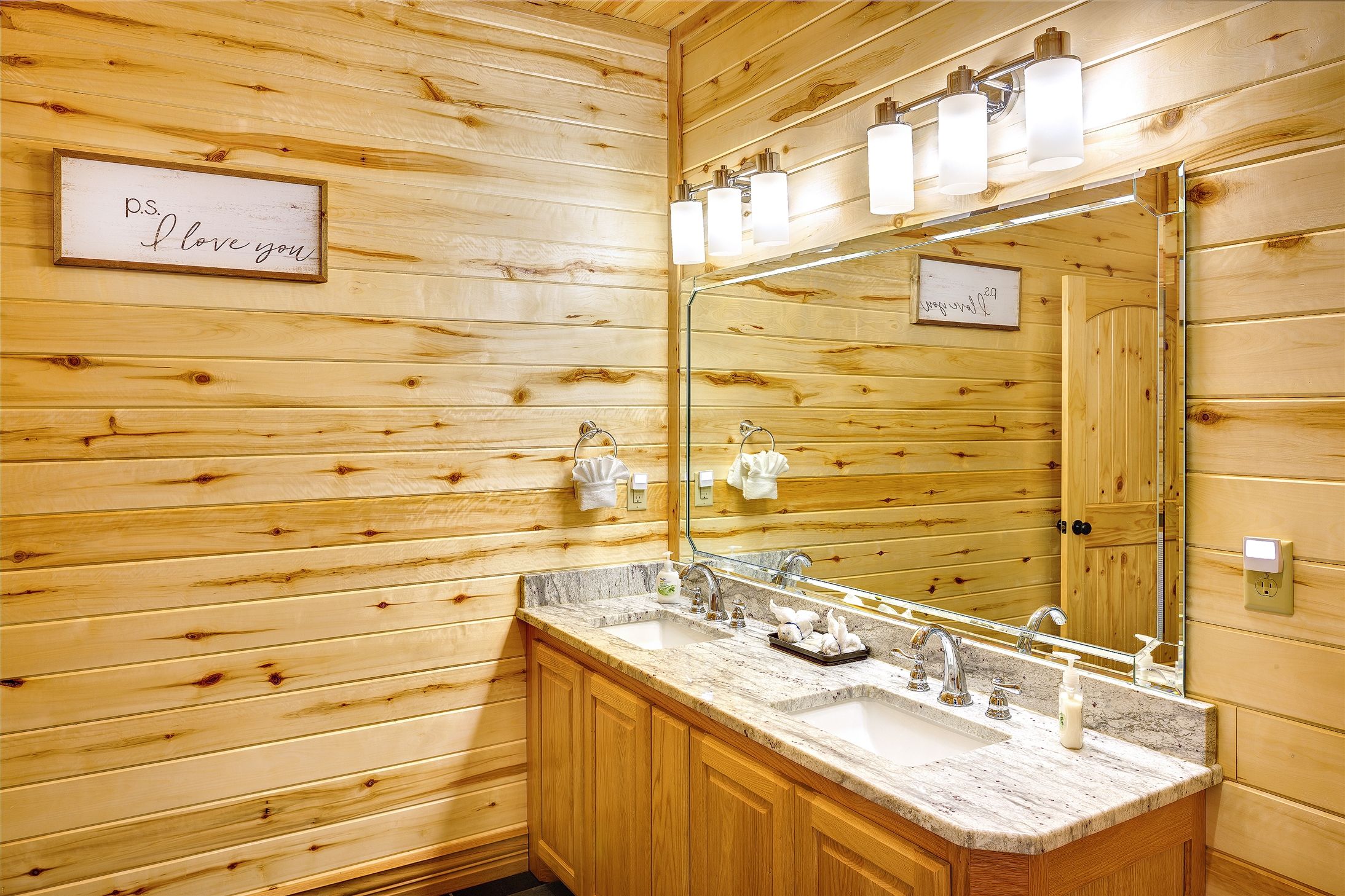 A wooden-paneled bathroom with a large mirror, two sinks, and a towel rack.