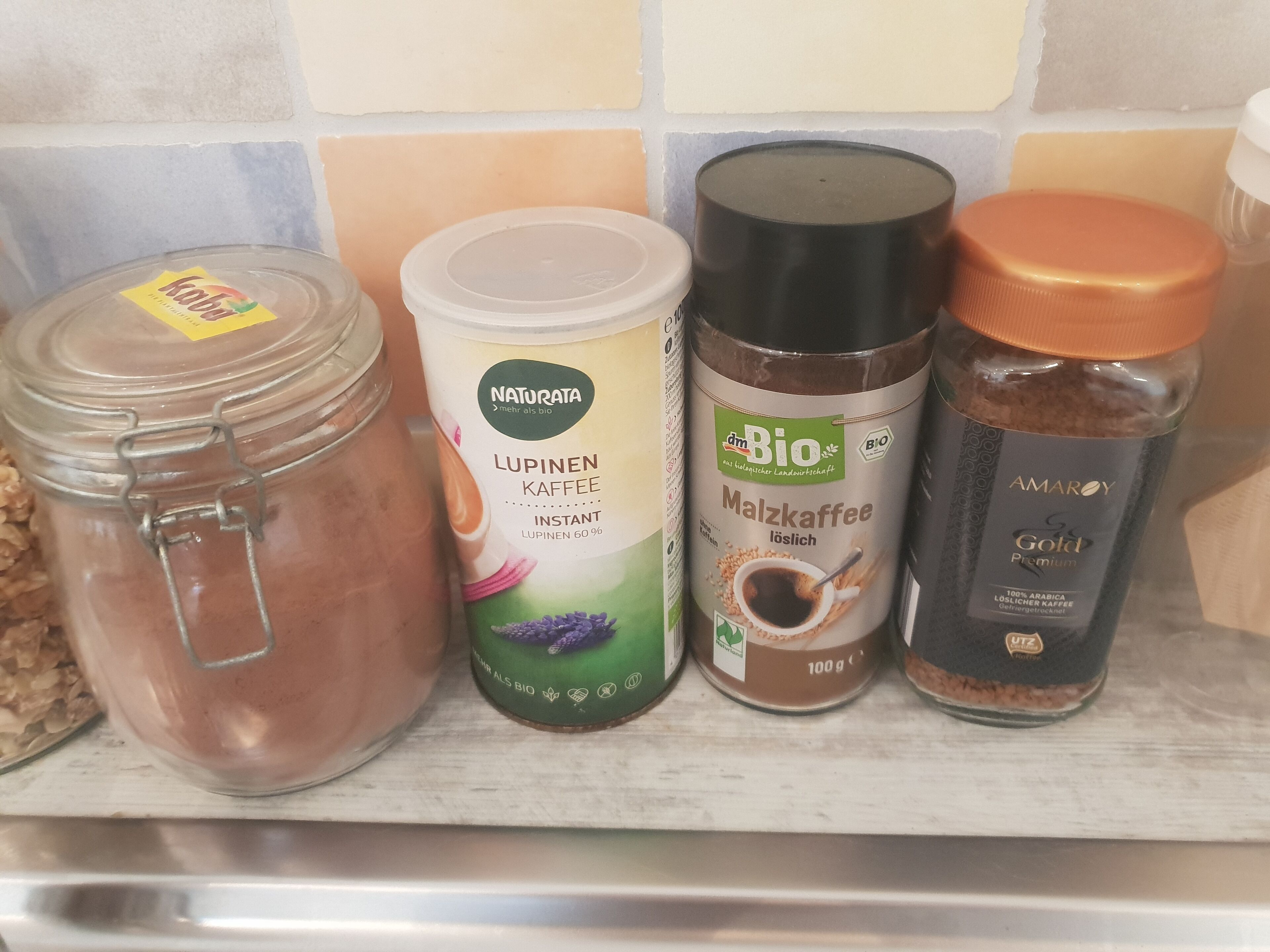 Ein Regal mit vier Behältern: ein Glasgefäß, ein weißer Behälter mit der Aufschrift 'Lupinen Kaffee', ein schwarzer Behälter mit der Aufschrift 'Bio Malzkaffee' und ein Gefäß mit einem orangefarbenen Deckel.