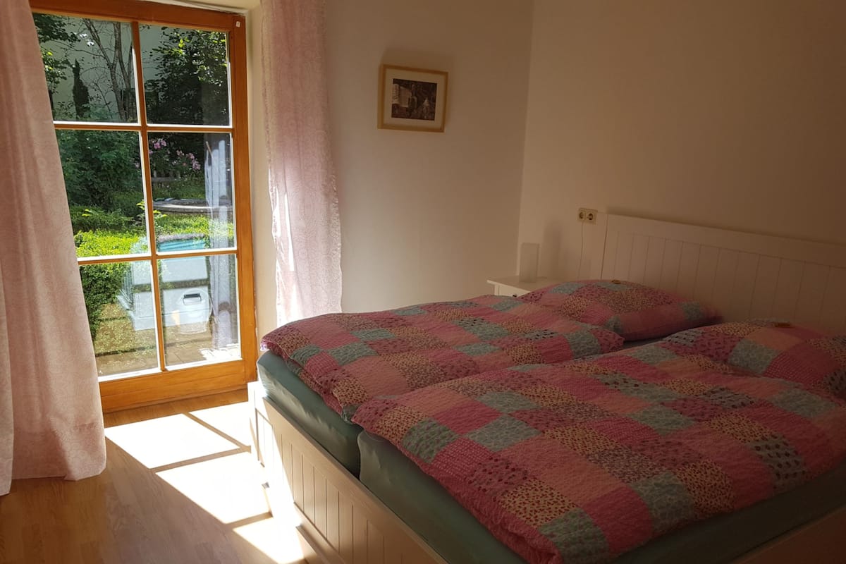 Ein Schlafzimmer mit großem Fenster, einem Bett mit gemustertem Überwurf und einem kleinen Bild in einem Rahmen an der Wand.
