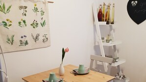 A dining area with a wooden table and chairs, a small vase with a pink flower, and a wall-mounted shelf with bottles and jars. - Martina's place (Rottenbuch)