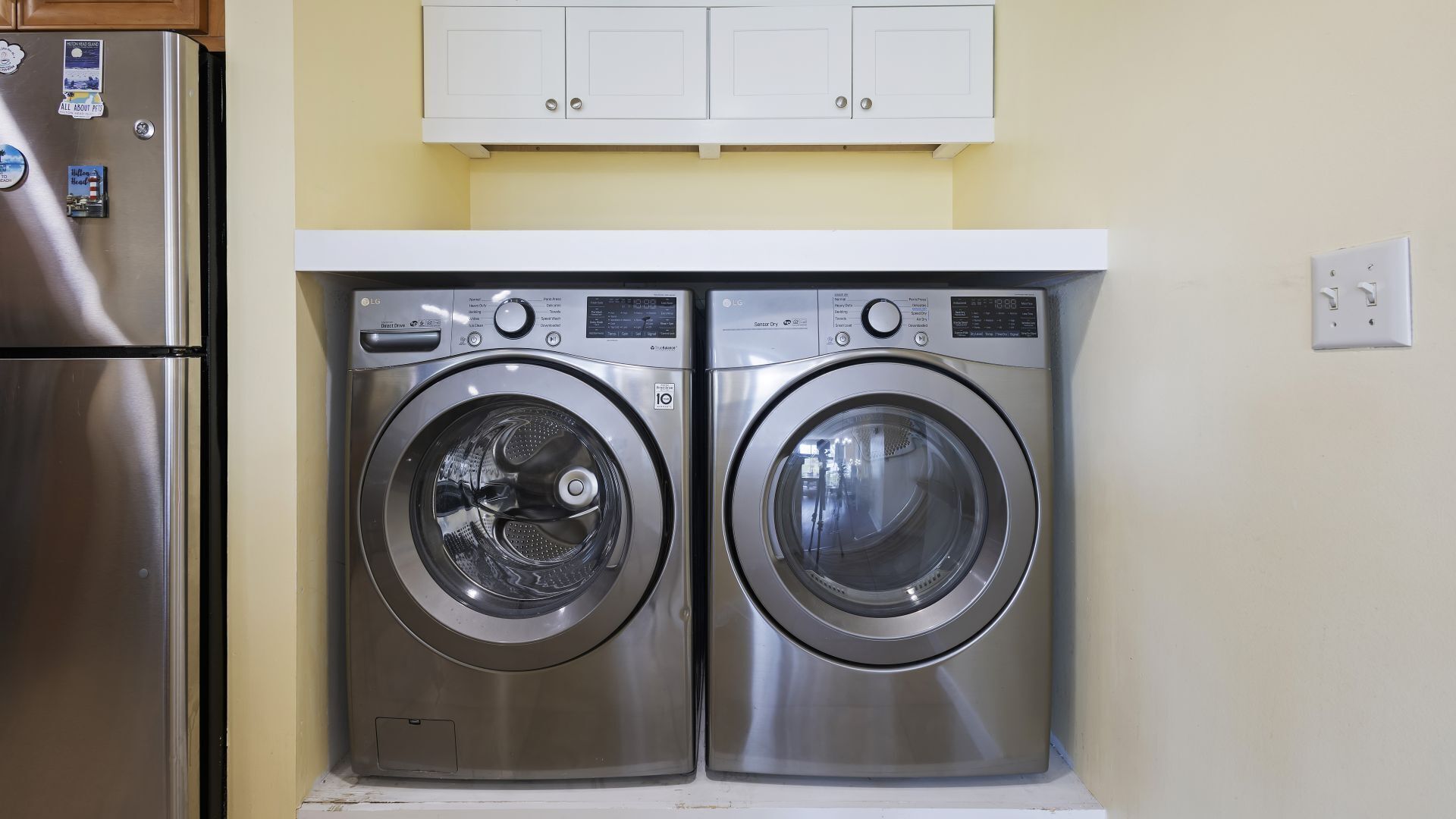 Un espace buanderie compact comprenant deux lave-linge LG à chargement frontal, un réfrigérateur en acier inoxydable et un meuble blanc placé au-dessus.