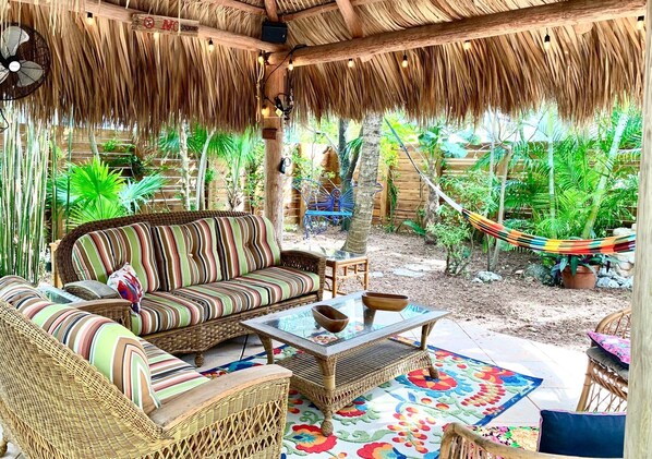 A covered outdoor seating area with wicker furniture, a glass-top coffee table, and a hammock.