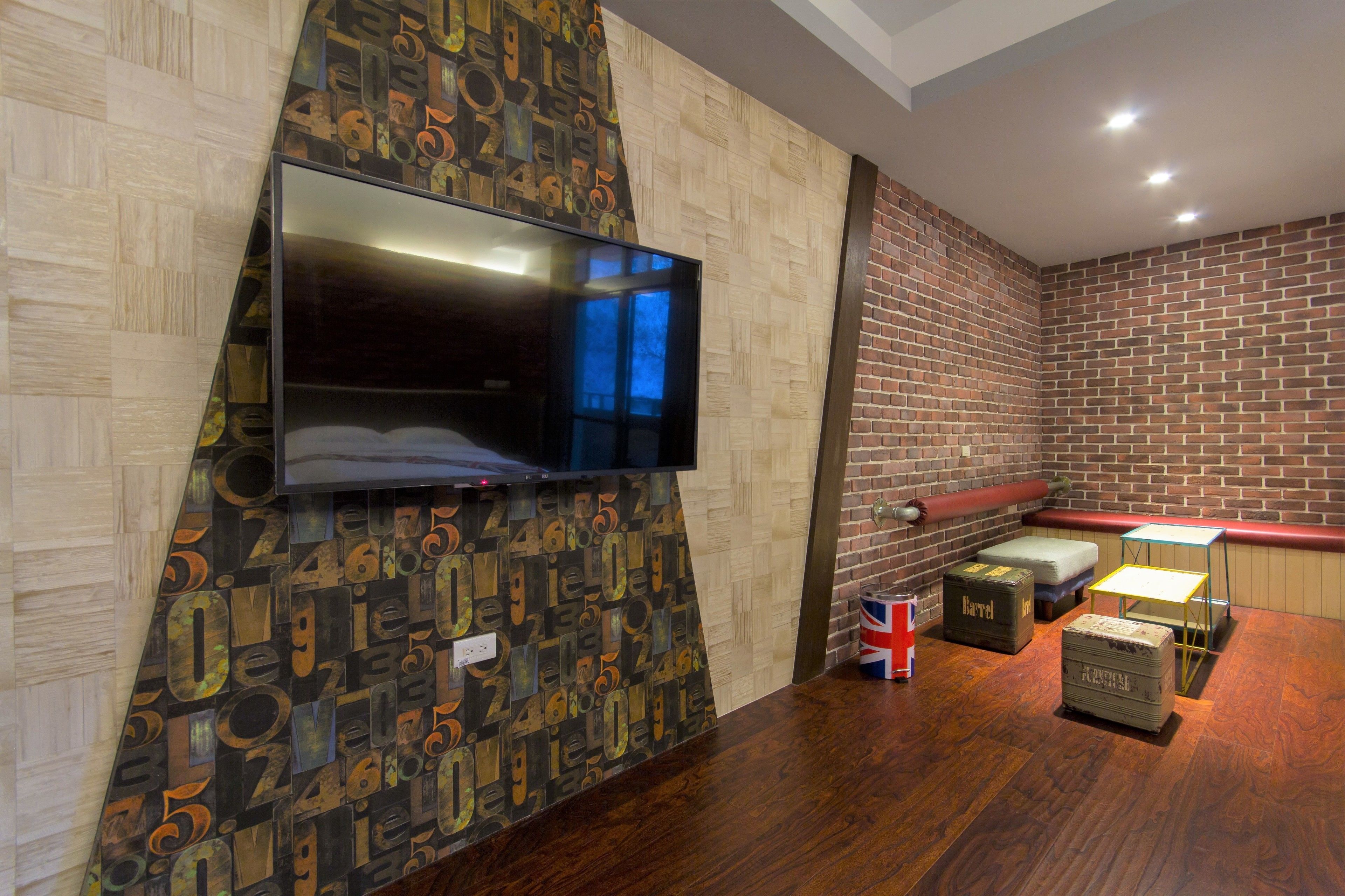 A modern room with a flat-screen TV mounted on a wall with a geometric pattern, a brick wall, a wooden bench, and a small table with a lamp.
