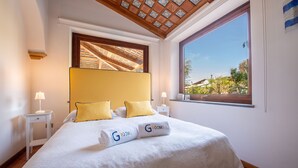 Une chambre d’hôtel avec un grand lit, deux tables de chevet avec des lampes, une fenêtre donnant sur un paysage verdoyant et un plafond orné de motifs.