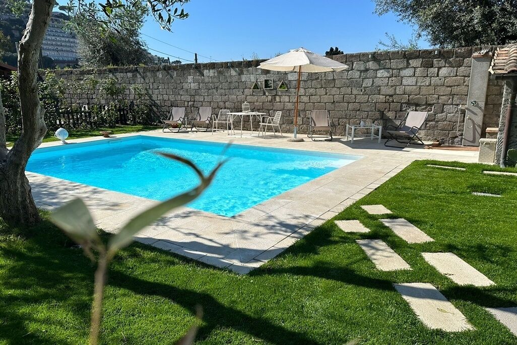 A swimming pool with a stone wall, surrounded by a grassy lawn and a few lounge chairs.