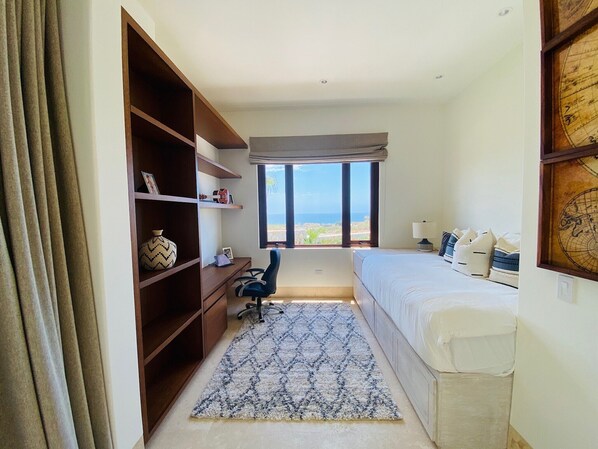 A bedroom with a bed, a desk, a chair, a bookshelf, and a large window with a view of the ocean.