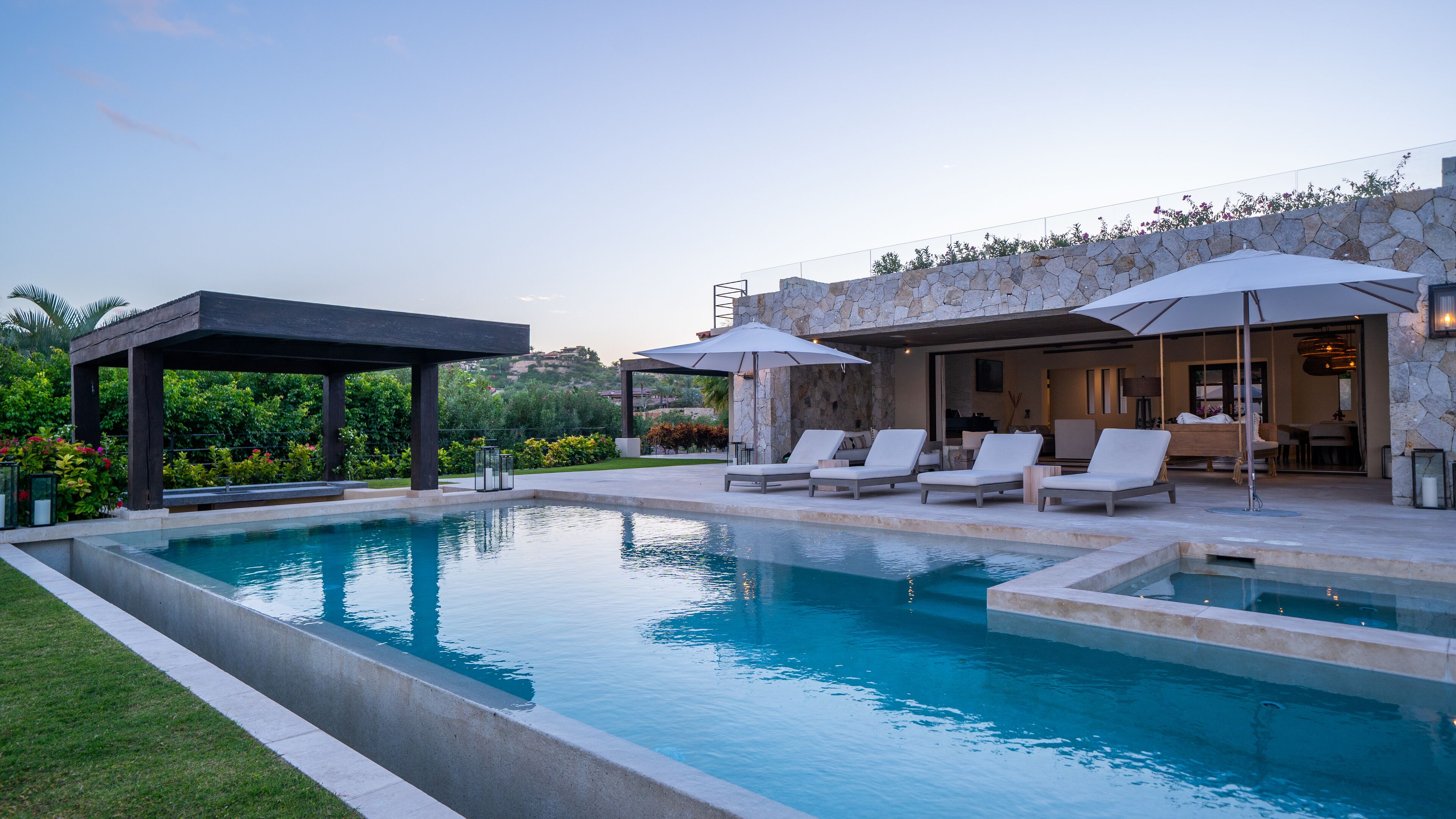 Una piscina con sillas de descanso y sombrillas, rodeada por un muro de piedra y vegetación.