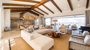 Amplia sala de estar con un sofá modular grande, un sillón estampado y una mesa de centro de madera. Se ve un piano al fondo, y la habitación tiene techos altos con vigas a la vista.