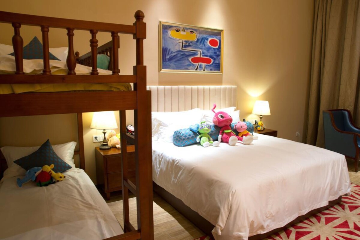 Une chambre d’hôtel avec un lit superposé, un lit simple et une table de chevet avec une lampe. Il y a un tableau au mur et une chaise dans un coin.