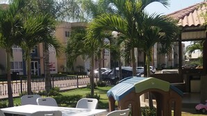 Area esterna con tavoli e sedie, circondata da palme e un piccolo parco giochi.