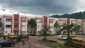 Un edificio a più piani con facciata rosso e beige, circondato da palme e un'area di parcheggio pavimentata.