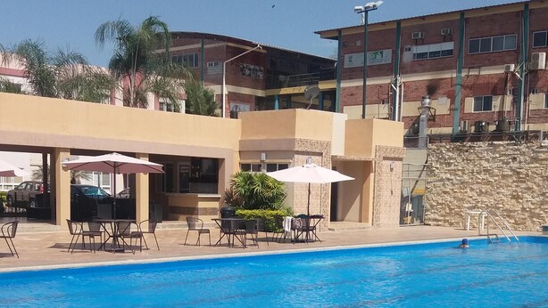 Una piscina con sedie a sdraio e ombrelloni, circondata da edifici e un muro in pietra, con una scala.