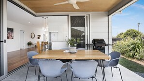 An open-plan living space with a wooden dining table, grey chairs, a ceiling fan, and a barbecue grill outside.