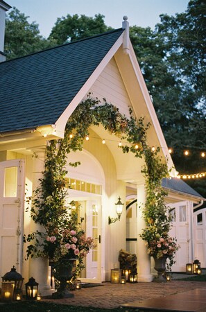 A house with a white facade, a dark roof, and a covered porch adorned with climbing plants and flowers. - Enjoy our 3-story, family-friendly Carriage House Fireplace and outdoor fire pit (West Redding)