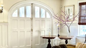 A room with a large arched white door, a wooden table with a vase of flowers, a sofa with a throw blanket, and a dresser with drawers. - Enjoy our 3-story, family-friendly Carriage House Fireplace and outdoor fire pit (West Redding)