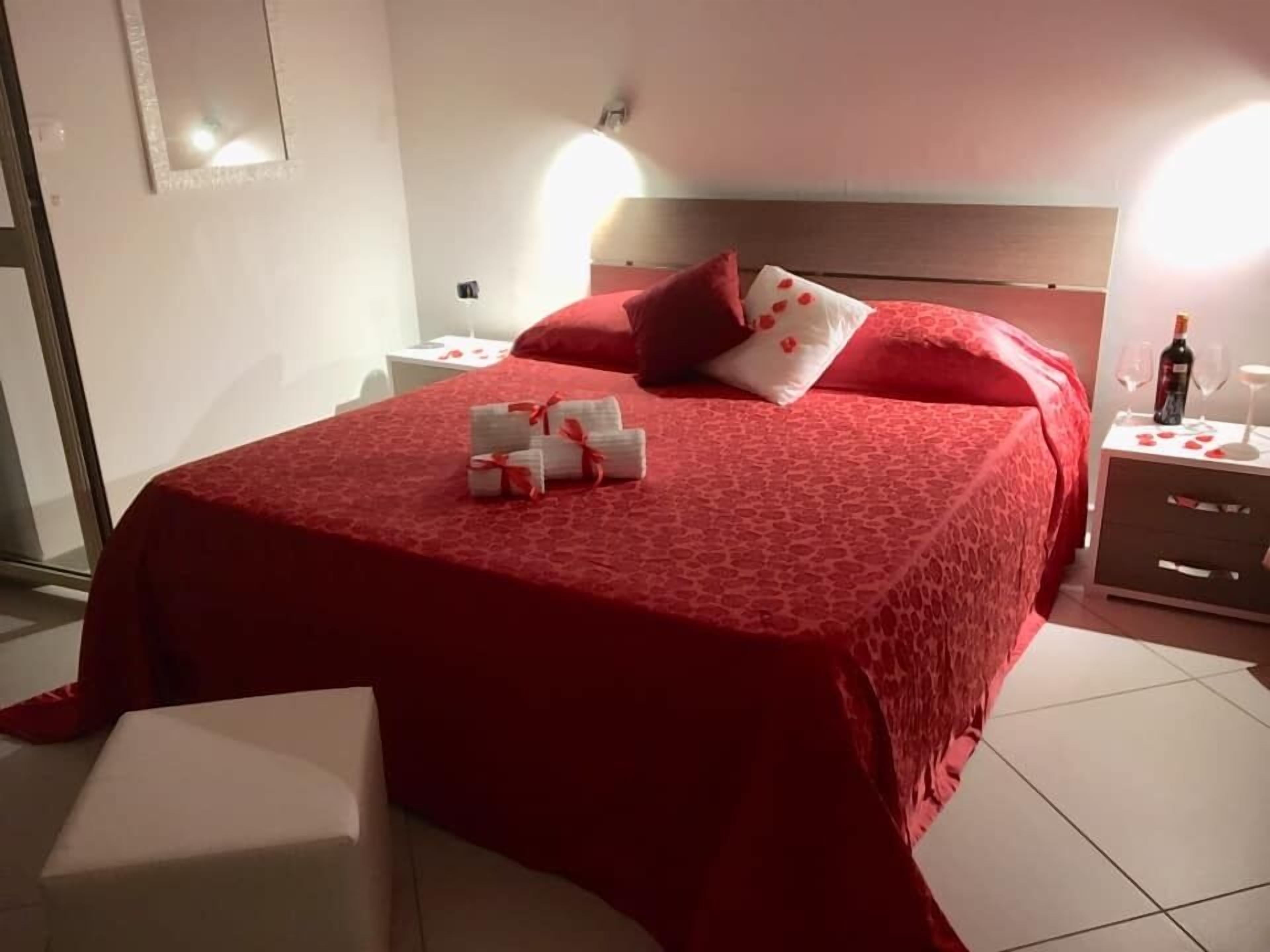 Un lit bien fait, recouvert d’une couverture rouge, avec un oreiller orné de motifs en forme de cœur rouge, et une table de chevet sur laquelle se trouvent une bouteille et des verres.