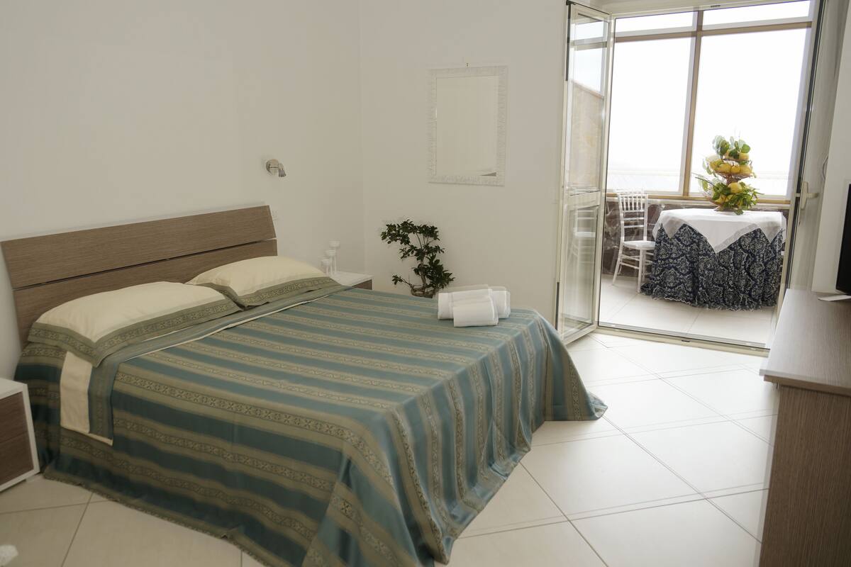 A bedroom with a bed, a bedside table, a plant, and a view of a balcony with a table and chairs.