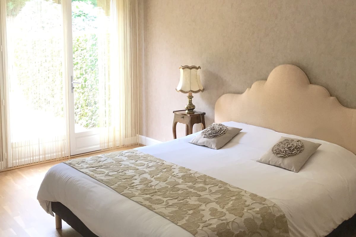 Un lit bien fait, recouvert d’une couvre-lit à motifs, agrémenté de deux coussins décoratifs et d’une lampe de chevet, et offrant une vue sur la verdure par la fenêtre.