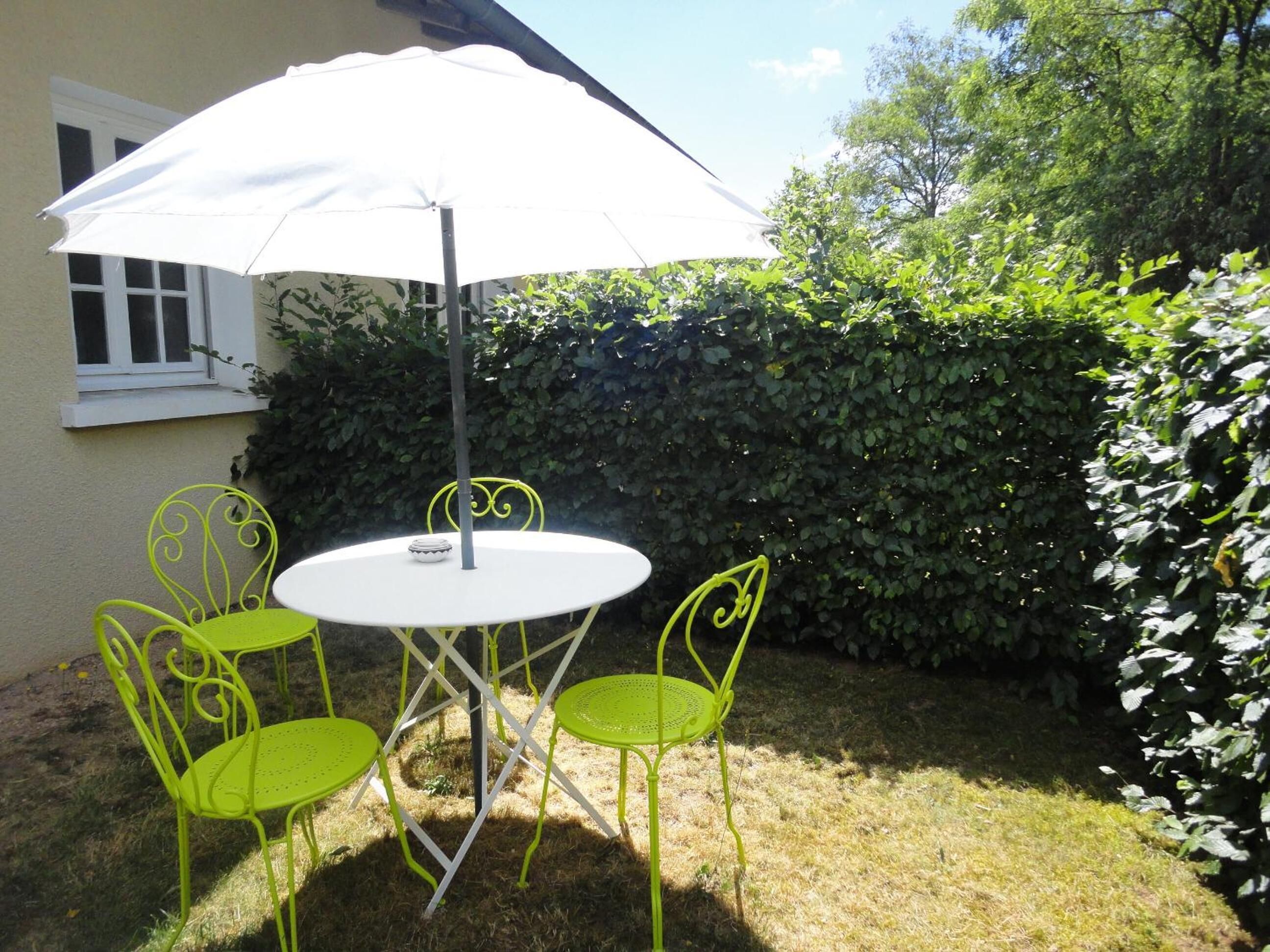 A round table with four green chairs and a white umbrella in a garden setting.