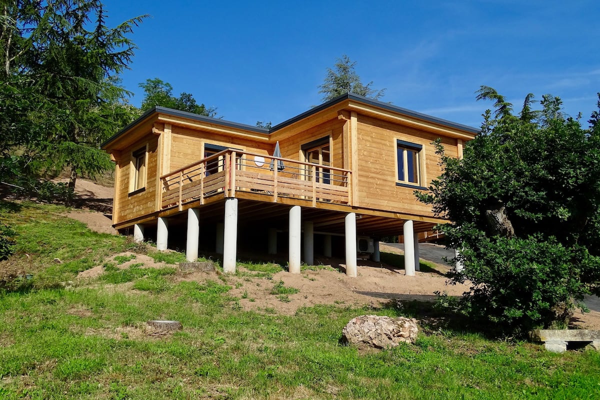 Une maison en bois à deux étages, dotée d’un balcon soutenu par des colonnes, entourée de verdure et d’arbres.