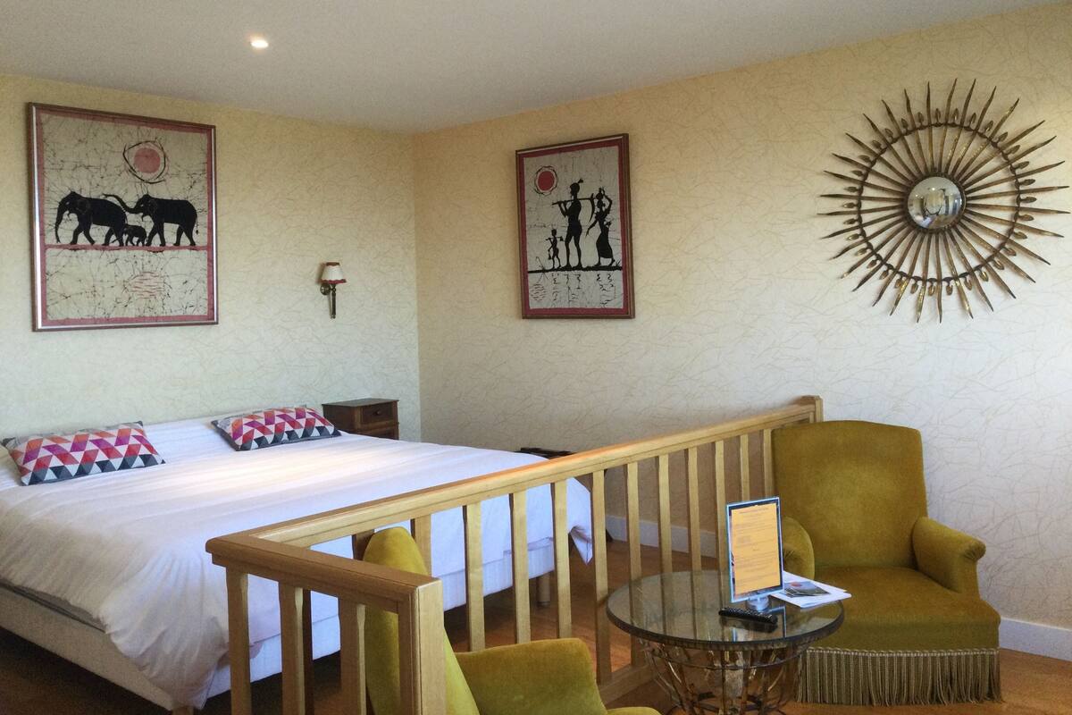 Une chambre d’hôtel avec un lit, un fauteuil vert, une petite table et des œuvres d’art murales.