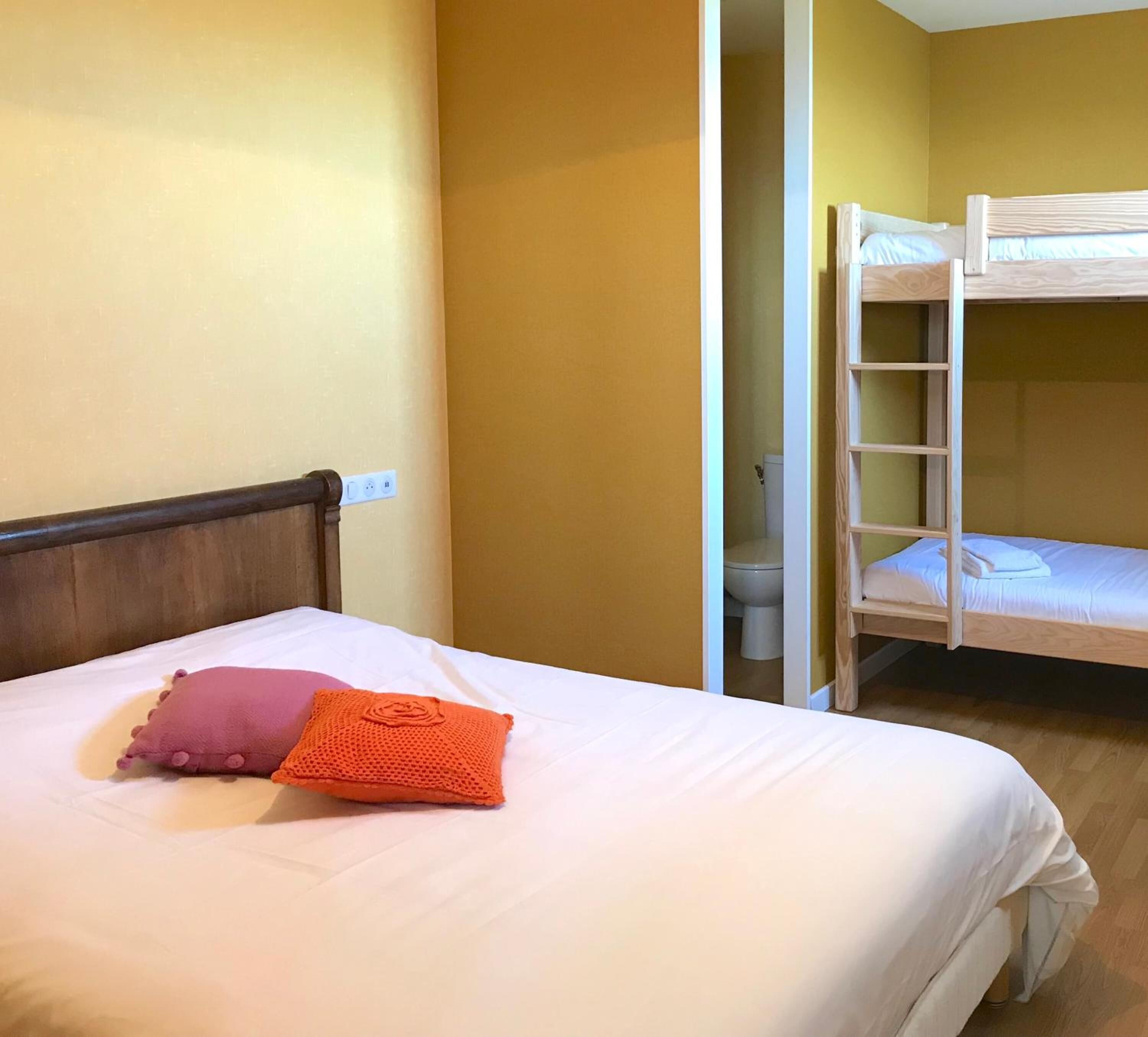 Una cama individual con cabecera de madera, almohada rosa y naranja, y una litera a la derecha.