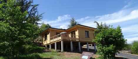 A two-story wooden house with a balcony, surrounded by greenery and a clear sky.