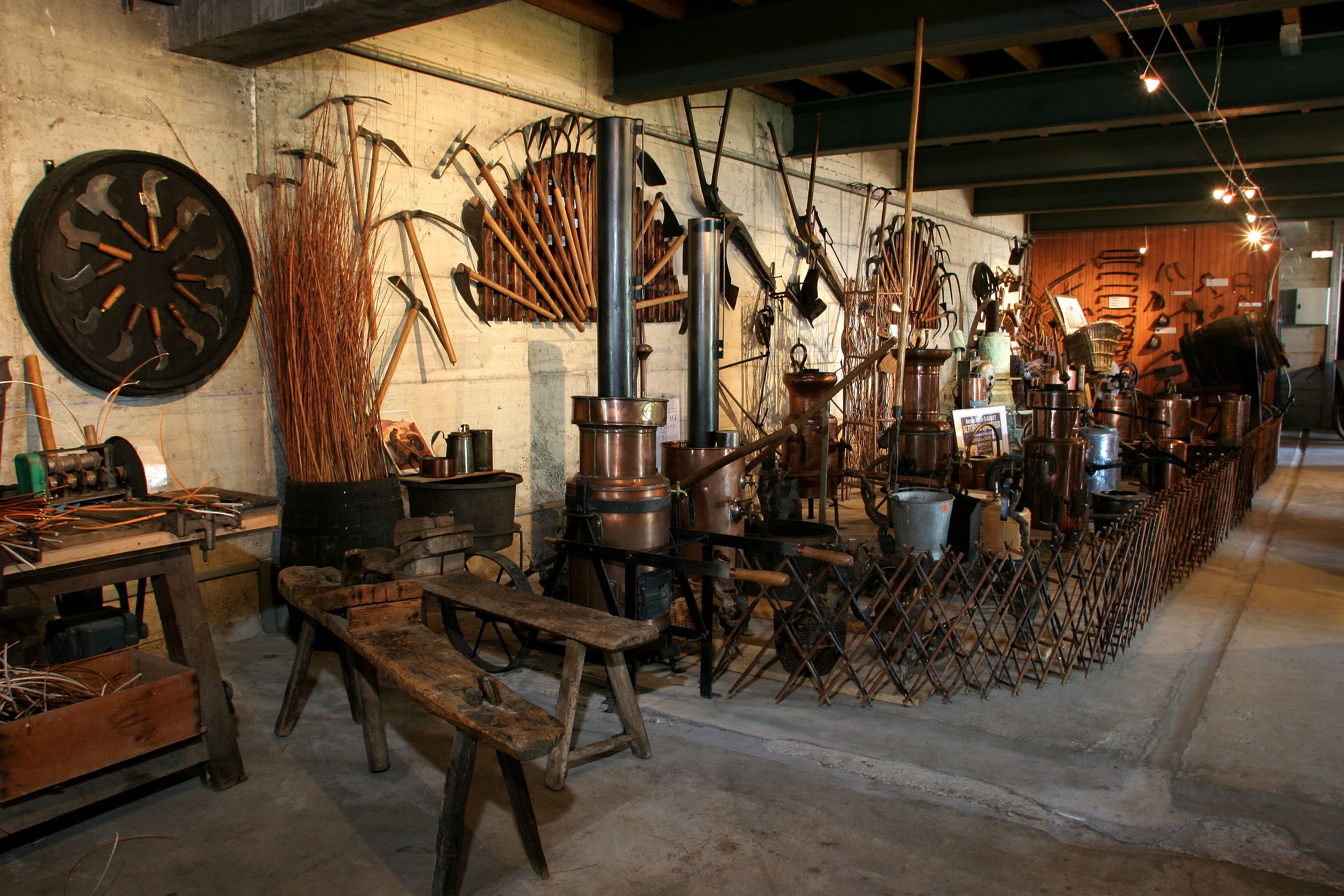 Un atelier rustique équipé de divers outils et matériels, notamment une grande scie circulaire, des établis en bois et des conteneurs métalliques.