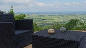 Un espacio de descanso al aire libre con mobiliario de mimbre y vistas a un paisaje verde y edificios a lo lejos.
