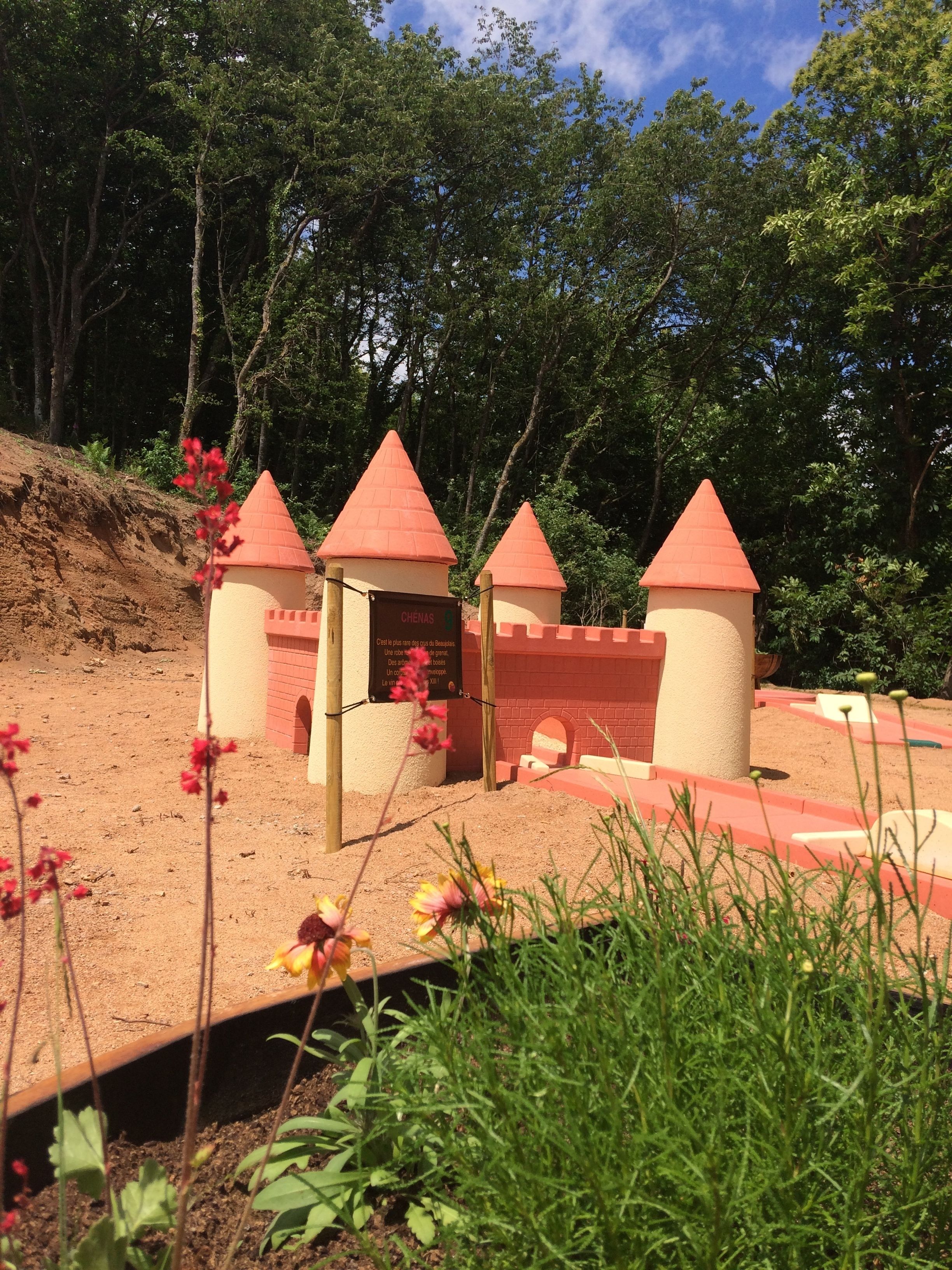A castle-like structure with three towers, surrounded by greenery and a dirt path.