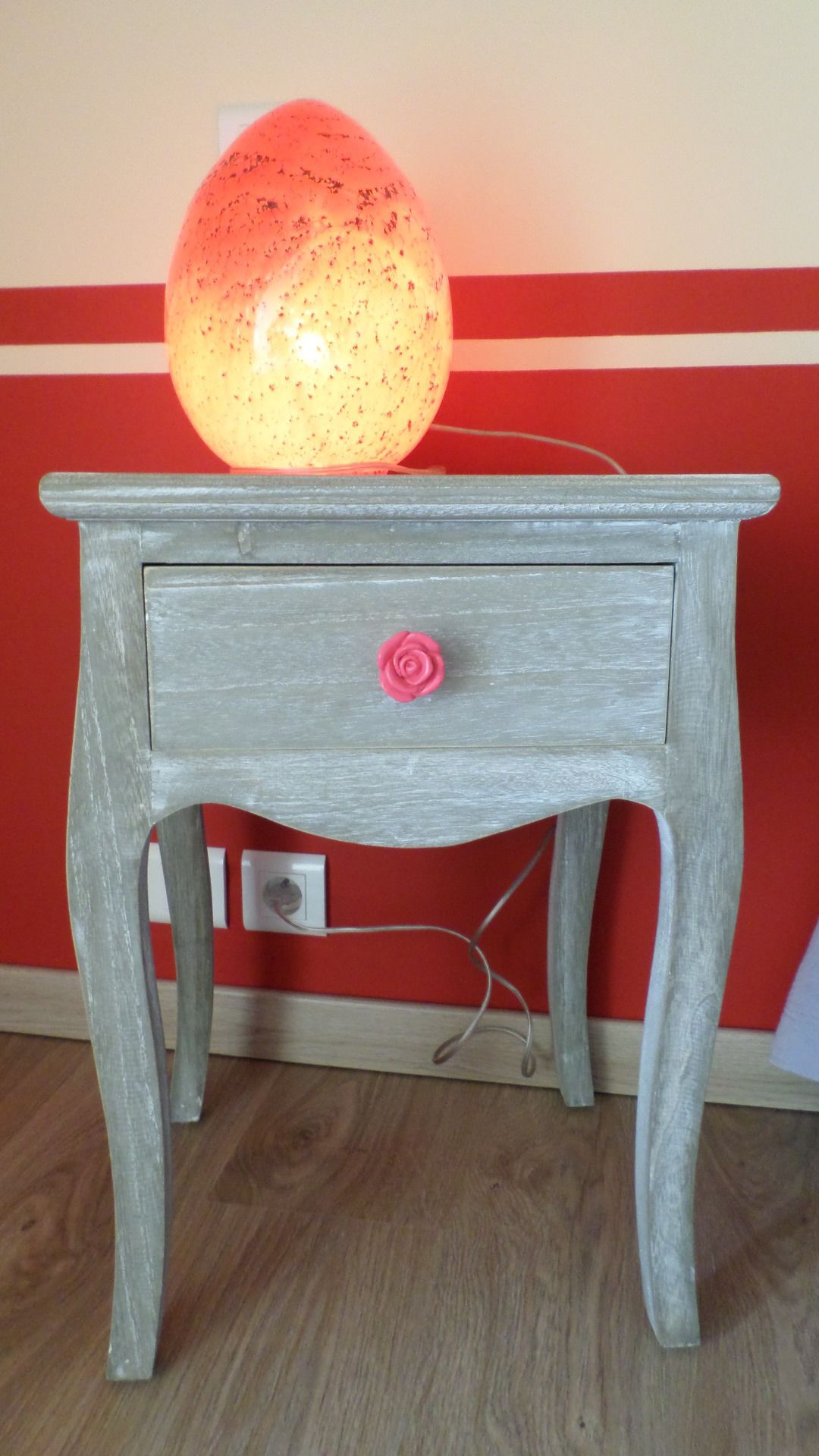 A wooden nightstand with a drawer, a pink rose decoration, and a lamp on top.