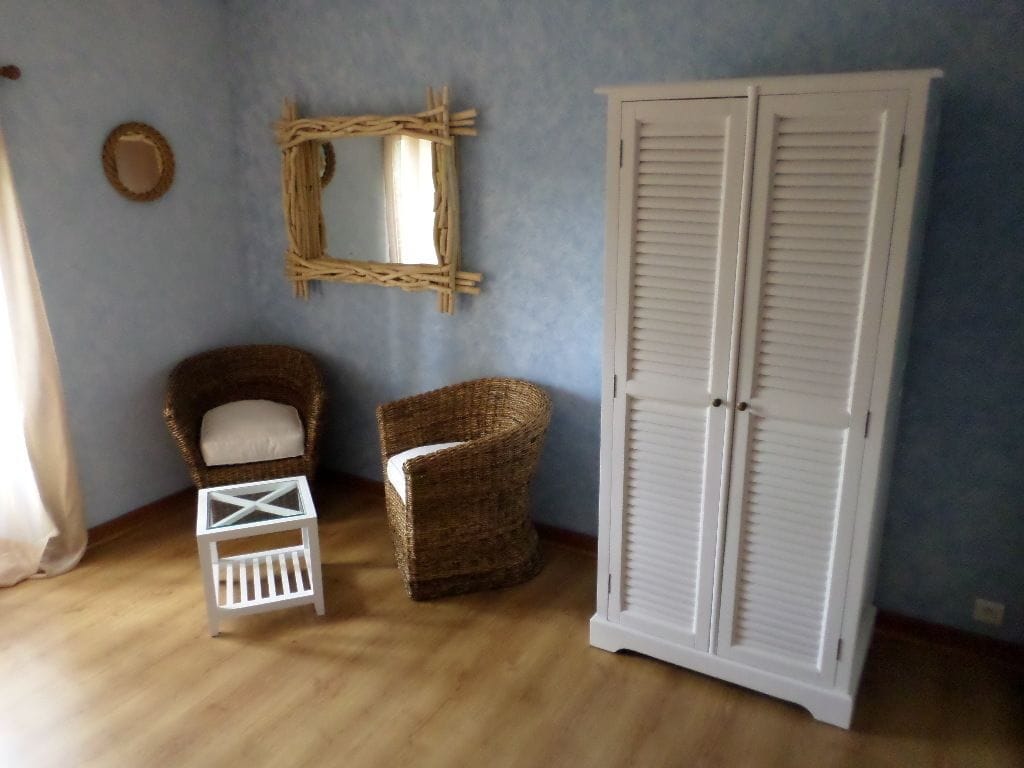Una habitación con una silla de mimbre, un taburete blanco pequeño, un armario blanco y un espejo tejido en la pared.