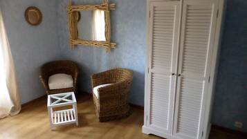 Una habitación con una silla de mimbre, un taburete blanco pequeño, un armario blanco y un espejo tejido en la pared.