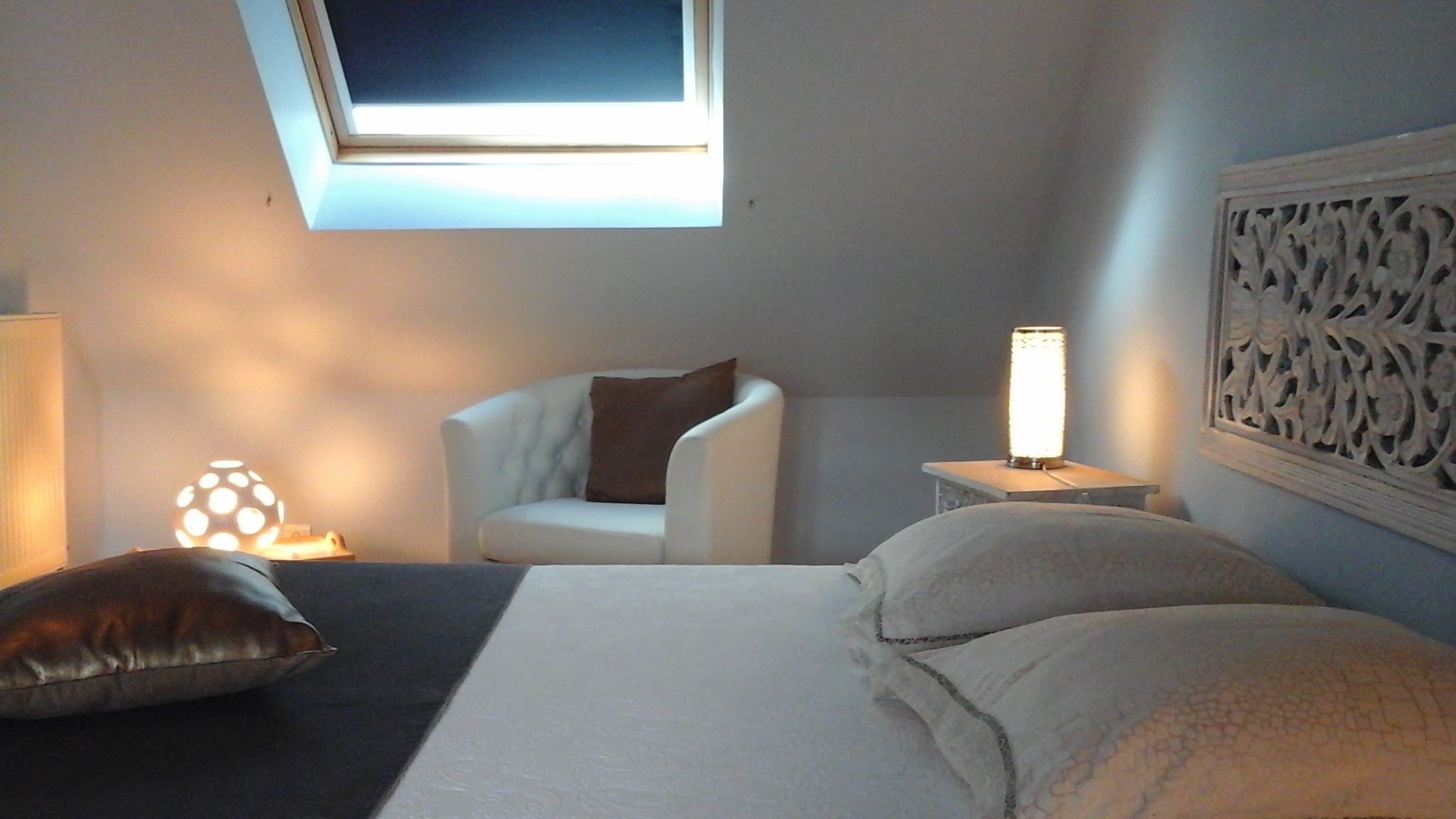 A bedroom with a bed, a chair, a nightstand, a wall-mounted light, and a skylight.