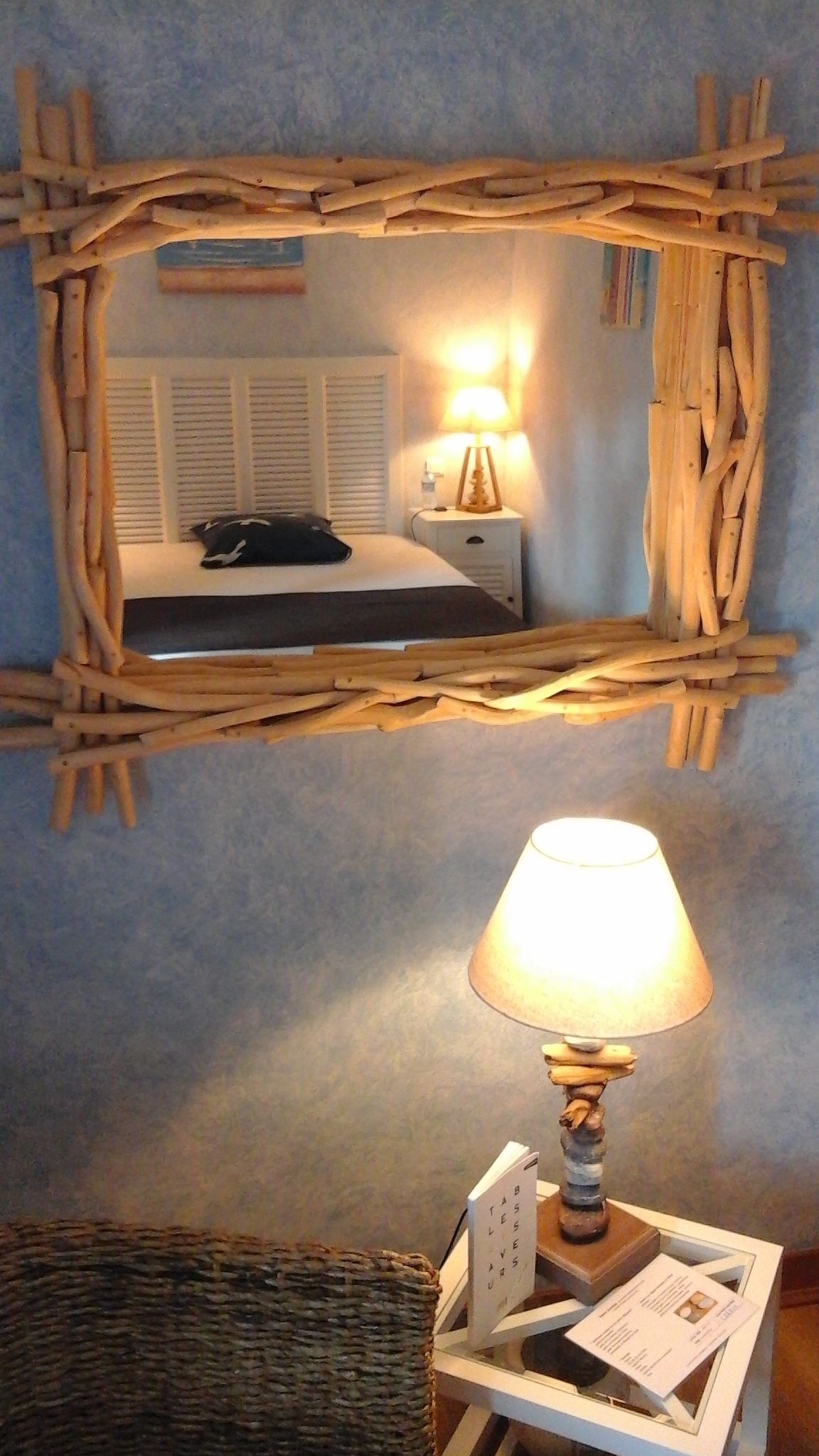 A room with a rustic wooden mirror on the wall, a bedside table with a lamp, a bed, and a wicker chair.