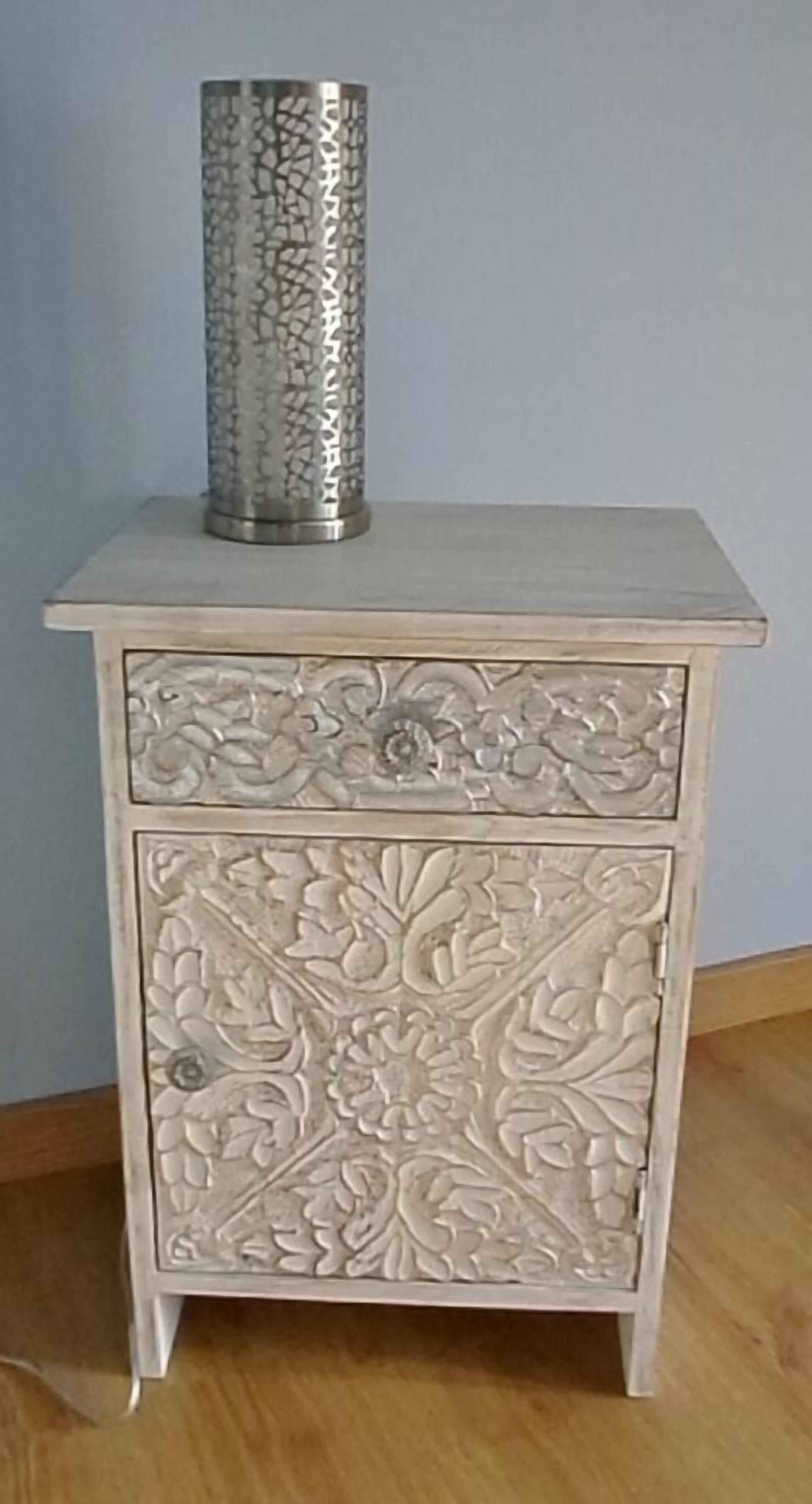 Een nachtkastje met witte lak en gedetailleerde bloemdecoraties, met een gestructureerd metalen object erboven.