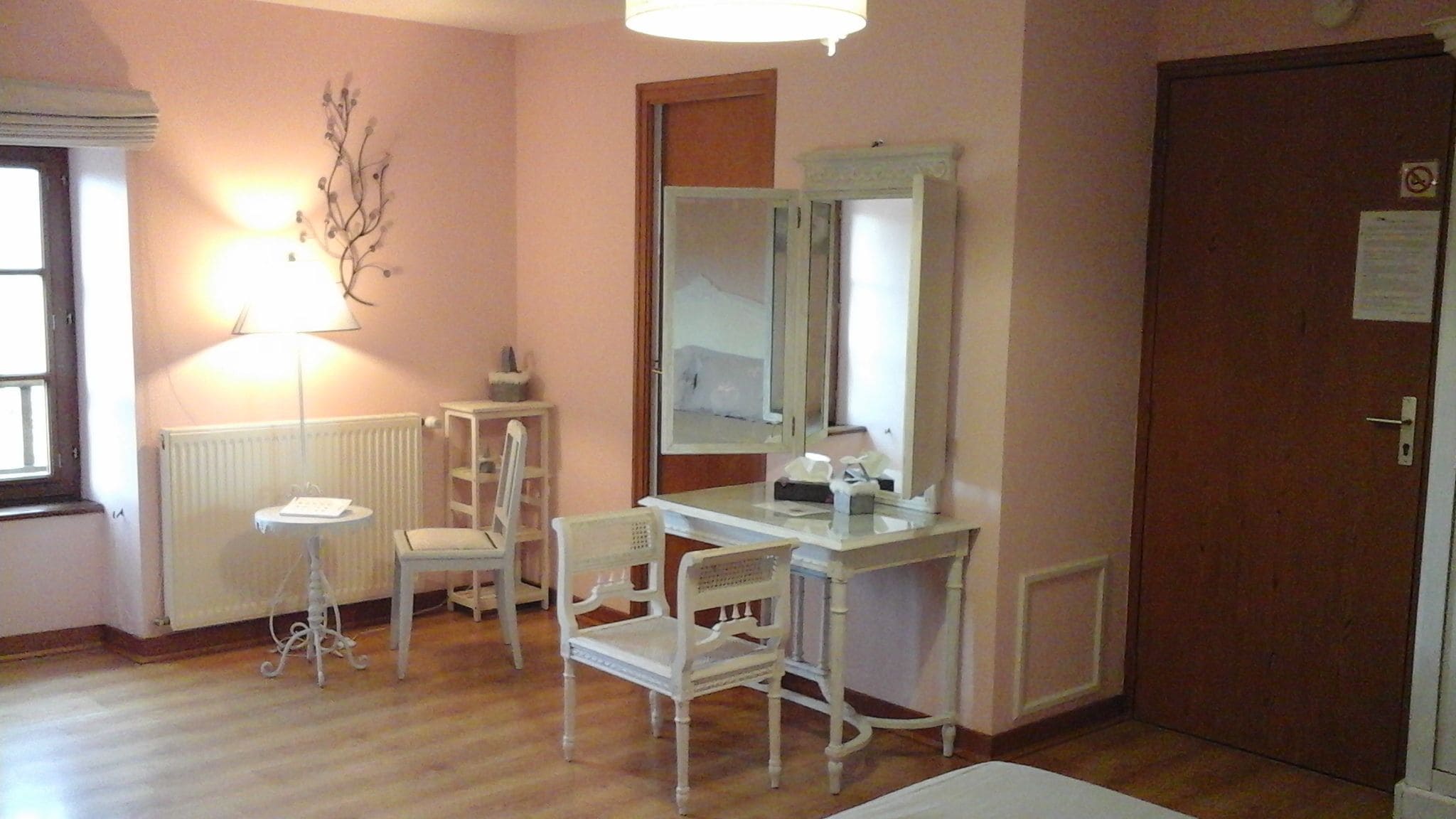Une pièce avec un plancher en bois, un bureau blanc avec un miroir, deux chaises blanches, une petite table avec une lampe et un radiateur.