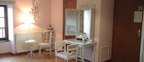 Une pièce avec un plancher en bois, un bureau blanc avec un miroir, deux chaises blanches, une petite table avec une lampe et un radiateur.