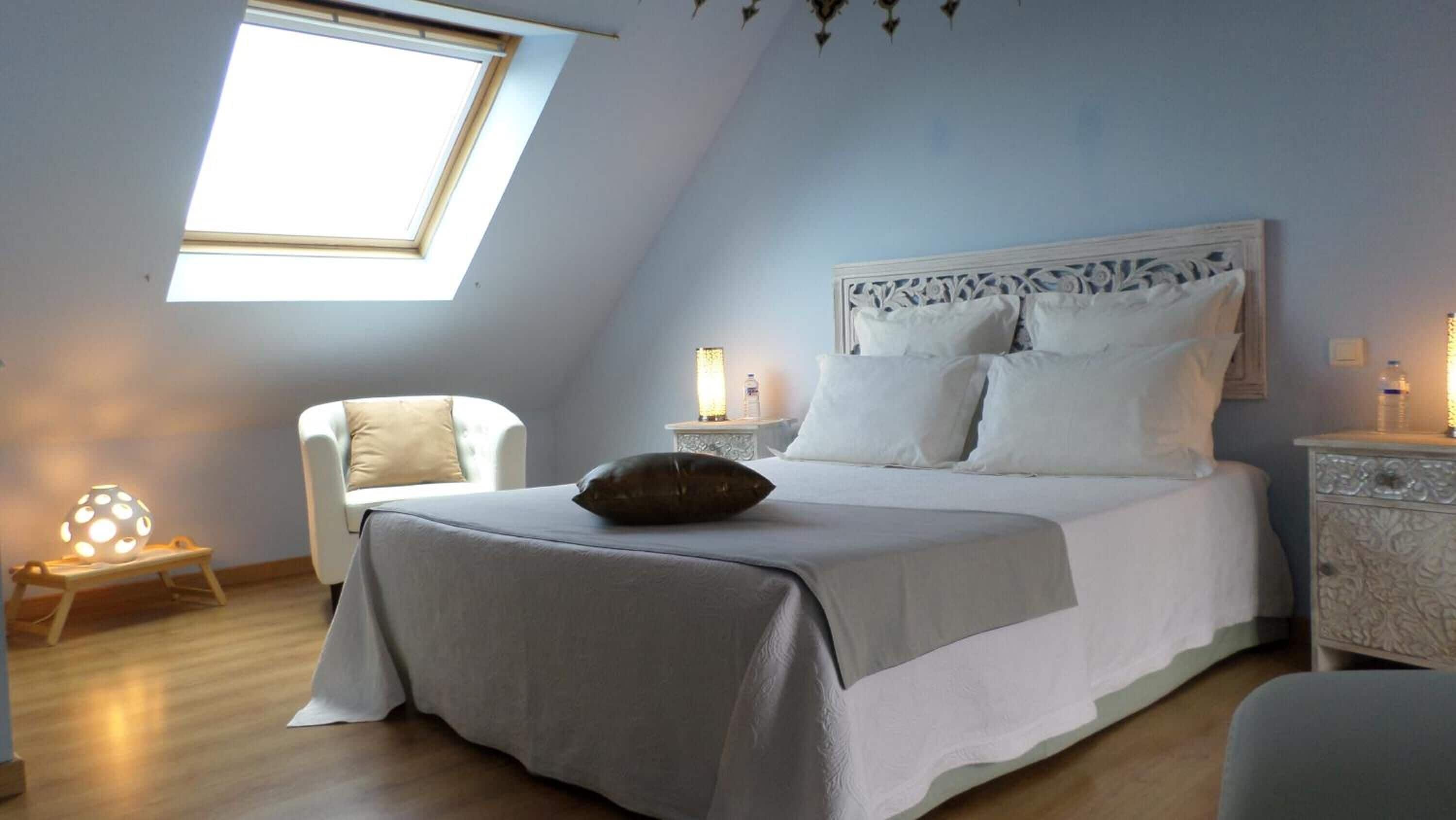 Un dormitorio con tragaluces, una cama con ropa blanca, una mesita de noche con lámpara y una silla.