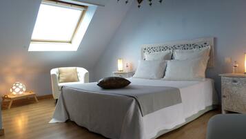 Un dormitorio con tragaluces, una cama con ropa blanca, una mesita de noche con lámpara y una silla.