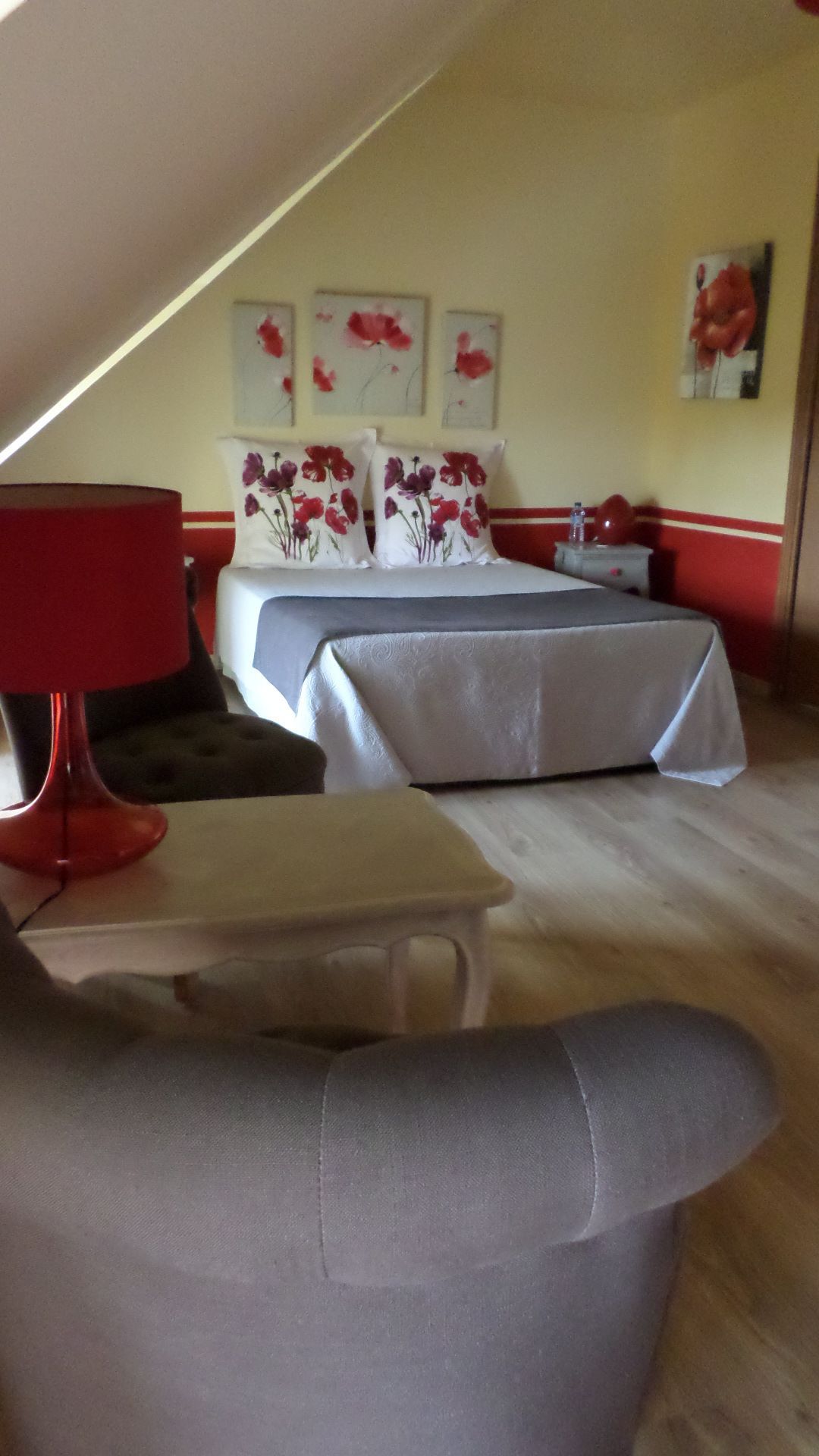 A hotel room with a bed, a chair, and a table. There are floral paintings on the wall.