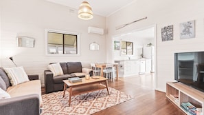 Uma sala de estar moderna com um sofá, mesa de centro e televisão. Há uma área de cozinha com mesa e cadeiras.