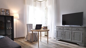 A modern living room with a sofa, a table with a laptop, a TV on a cabinet, and a bookshelf.
