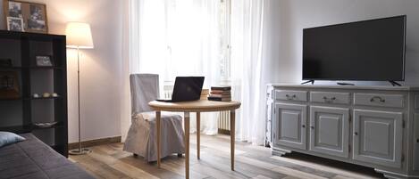 A modern living room with a sofa, a table with a laptop, a TV on a cabinet, and a bookshelf.