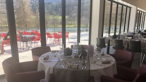A restaurant with round tables set for dining, large windows offering a view of a lake and trees, and red chairs outside. - Base de Loisirs du Lac de la Moselotte (Saulxures-sur-Moselotte)