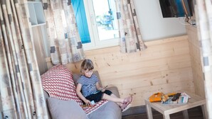 A cozy room with a wooden wall, a window with blue curtains, a television mounted on the wall, and a small table with a magazine and a remote control. - Base de Loisirs du Lac de la Moselotte (Saulxures-sur-Moselotte)