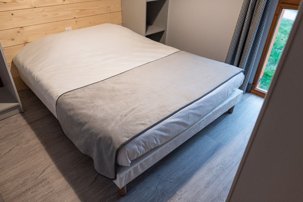 Una cama individual con colchón gris y ropa de cama blanca en una habitación con paneles de madera y una ventana con cortinas.